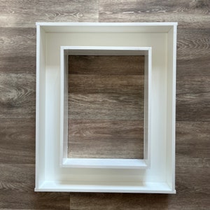 May include: Two white picture frames, one nested inside the other, on a woodgrain background.