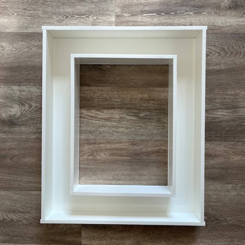 May include: Two white picture frames, one nested inside the other, on a woodgrain background.