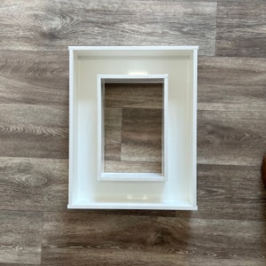 May include: Two white square frames, one nested inside the other. The outer frame is slightly larger than the inner frame.