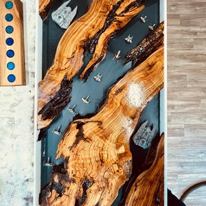 Pode incluir: Mesa de centro retangular com moldura branca. A mesa apresenta um design de rio em resina epóxi cinza escuro com naves espaciais em miniatura e elementos de madeira embutidos. A madeira tem um grão natural e uma cor marrom quente.