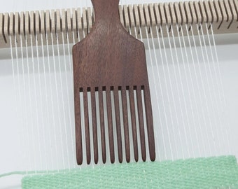 Könnte beinhalten: Ein Holzkamm mit abgerundetem Griff, der zum Weben verwendet wird. Der Kamm liegt auf einem Webstuhl mit weißem Garn und einem grünen gewebten Streifen.