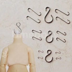 Puede incluir: Una colección de pequeños ganchos metálicos plateados de varias formas. Algunos tienen forma de S, mientras que otros son rectos con extremos curvos. Los ganchos se muestran sobre una superficie de color claro, con un torso de muñeca en primer plano.