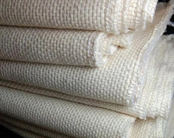 May include: Close-up of a stack of off-white woven fabric. The fabric has a textured, grid-like pattern and appears to be made of a natural fiber. The edges of the fabric are slightly frayed, suggesting a raw or unfinished quality. The fabric is folded and stacked.