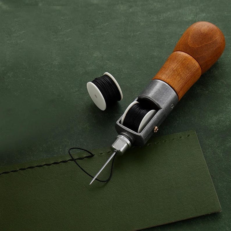 May include: A close-up of a leather sewing awl with a wooden handle and a metal needle. The awl is threaded with black thread and is resting on a green surface. There is a spool of black thread to the left of the awl.