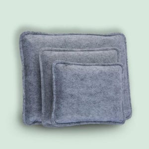 May include: Three gray felt pillows of varying sizes are stacked together. The pillows have a rectangular shape with rounded corners and a stitched border. The felt material has a soft, textured appearance.
