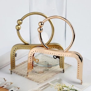 Op de afbeelding: Twee metalen portemonneeframes in goud en roségoud. Elk frame heeft een afgeronde handgreep met decoratieve balaccenten. De frames hebben een rechthoekige basis met kleine gaatjes om te naaien. Het roségouden frame staat voor het gouden frame.