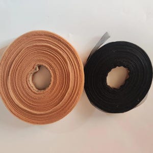 Op de afbeelding: Twee rollen lint worden getoond. Eén is lichtbruin, de andere zwart. Beide zijn cirkelvormig met een gat in het midden. Het lichtbruine lint heeft een getextureerde uitstraling, terwijl het zwarte lint van een transparante stof lijkt te zijn.