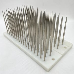 May include: A rectangular white base with numerous thin, silver metal needles protruding upwards. The needles are evenly spaced and appear sharp. The base has two circular holes. This item is likely a tool or instrument.