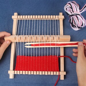 May include: A wooden loom with red yarn woven into a red and white pattern. The loom has a wooden shuttle and a wooden needle.