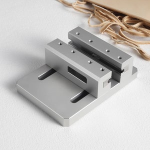 May include: A silver metal clamp with a rectangular base and two parallel, raised bars, each with screw holes. Light tan leather strips and a piece of leather are in the background. This tool is likely for crafting or woodworking.