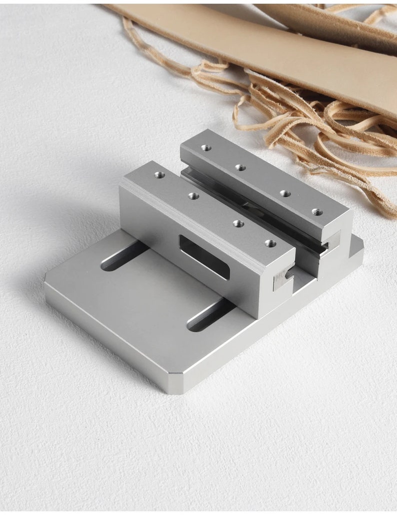 May include: A silver metal clamp with a rectangular base and two parallel, raised bars, each with screw holes. Light tan leather strips and a piece of leather are in the background. This tool is likely for crafting or woodworking.