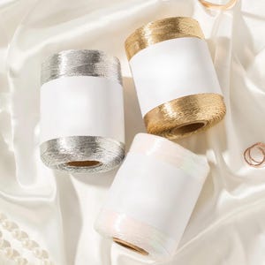 May include: Three cylindrical spools of metallic thread, one in silver, one in gold, and one in an iridescent white, each featuring a wide, blank white paper label wrapped around the center of the spool, arranged on a soft, draped fabric surface.