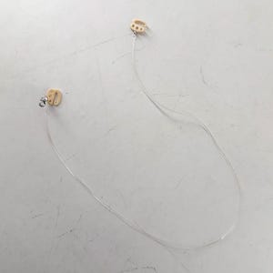 Puede incluir: Un par de audífonos beige conectados por un cable delgado y transparente. Los audífonos tienen una forma curva y están diseñados para colocarse detrás de la oreja. El cable es flexible y parece ser de plástico.