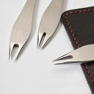 Puede incluir: Primer plano de varias herramientas plateadas para trabajar el cuero con un diseño bifurcado. Las herramientas se colocan sobre una superficie blanca junto a una pieza de cuero negro con costuras rojas. Las herramientas tienen un extremo afilado y puntiagudo.
