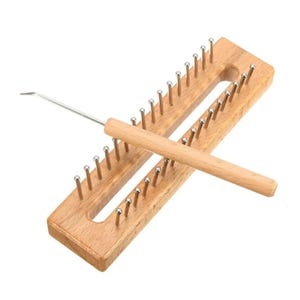 Puede incluir: Un telar de punto de madera con clavijas metálicas y un mango de madera. Un ganchillo de metal descansa sobre el telar. El telar es rectangular con un recorte central. Las clavijas están espaciadas uniformemente.