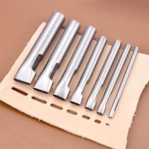 Puede incluir: Un conjunto de siete herramientas de trabajo de cuero de metal plateado, que incluyen punzones de diferentes tamaños, dispuestos sobre un trozo de cuero marrón claro. Las herramientas están diseñadas para perforar agujeros en cuero.