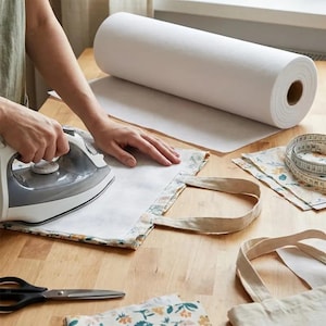 May include: A white iron is being used to press a light-coloured fabric tote bag on a wooden surface. A large roll of white interfacing material, a measuring tape, and black scissors are placed nearby, along with additional pieces of floral fabric.
