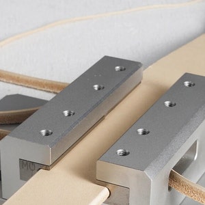 May include: A close-up shot of a leatherworking tool. Two silver metal clamps secure a light tan leather strip. Each clamp features several screw holes. A second, thinner leather strip is visible in the background.