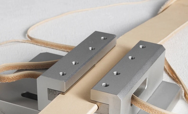 May include: A close-up shot of a leatherworking tool. Two silver metal clamps secure a light tan leather strip. Each clamp features several screw holes. A second, thinner leather strip is visible in the background.