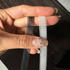 May include: Two narrow strips of mesh ribbon, one in black and one in white, featuring a consistent hexagonal honeycomb pattern throughout the length of the material. The ribbons are thin, flexible, and display a semi-sheer, open-weave texture.