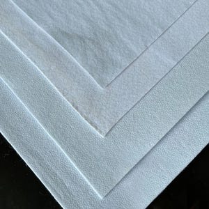 May include: A stack of light blue fabric sheets with varying textures. The sheets are arranged in a layered pattern, with the top sheet having a smooth surface and the lower sheets showing a textured, dotted pattern. The edges of the sheets are visible, creating a geometric design.