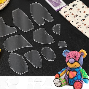May include: Clear acrylic bear pattern pieces with dashed stitch lines are arranged on a black surface. A colourful patchwork bear is in the lower right corner. A patterned fabric and a tag are also visible.