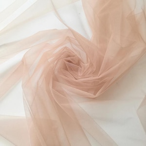 May include: A close-up of a swirl of sheer, blush-colored tulle fabric. The fabric is layered and creates a soft, delicate texture. The tulle is semi-transparent, allowing light to pass through, and is ideal for crafting and design projects.