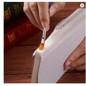 Puede incluir: Primer plano de la restauración de un libro. Un pincel con cerdas naranjas aplica pegamento al borde del libro. El lomo del libro está cubierto con tela blanca. El fondo incluye otros libros y una superficie de madera.