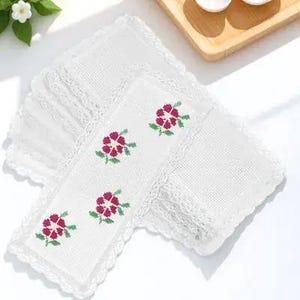 May include: A stack of white embroidered coasters with a floral design. Each coaster features a delicate lace trim and a cross-stitched pattern of red flowers with green leaves. The coasters are arranged on a white surface.