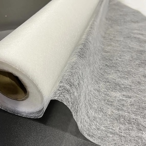 May include: A roll of white, semi-transparent, non-woven interfacing fabric with a fibrous, mesh-like texture. The material is wound around a cylindrical cardboard core, showing a dense, layered structure that is lightweight and thin.
