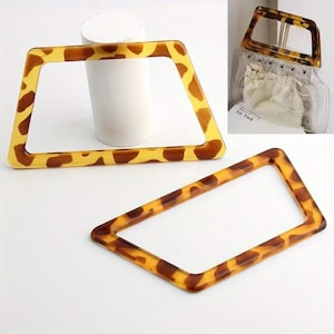 Pode incluir: Duas alças retangulares para bolsas com padrão de tartaruga. As alças são amarelo claro com manchas marrons. Uma alça está em primeiro plano e a outra no fundo. Uma bolsa transparente com alça está no fundo.