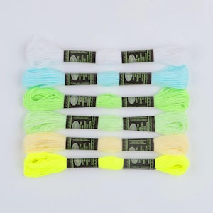 May include: Six skeins of glow-in-the-dark embroidery floss arranged horizontally. The colors from top to bottom are white, light blue, light green, lime green, pale yellow, and neon yellow. Each skein features a black paper label.