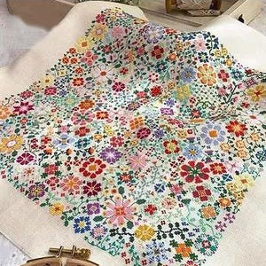 Könnte beinhalten: Ein farbenfrohes Kreuzstich-Blumenmuster auf weißem Stoff. Das Design zeigt eine Vielzahl von Blumen in Rot-, Gelb-, Blau-, Lila- und Orangetönen mit grünen Blättern. Der Stoff ist über einen hölzernen Stickrahmen gespannt.