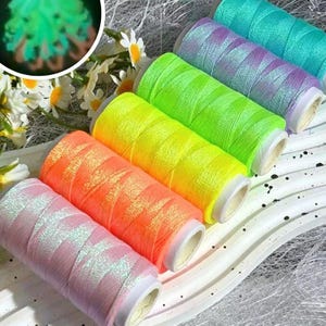 May include: A set of five cylindrical spools of thread in vibrant neon colours including pink, orange, yellow, green, and a purple-blue gradient. The thread features a reflective, shimmering finish and is arranged in a row next to white flowers.