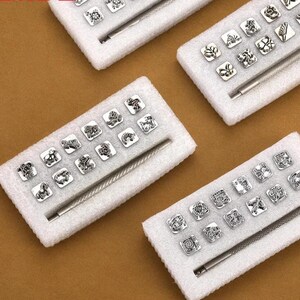以下が含まれることがあります： 様々なデザインの銀色の金属スタンプのコレクション。花柄や幾何学模様など、白いフォームトレイに配置されています。各トレイには金属製のツールが含まれています。スタンプは茶色の表面に表示されています。