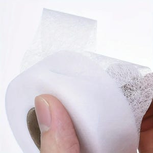May include: A roll of white, semi-transparent adhesive tape featuring a fine, fibrous, and web-like texture throughout the material. The tape is wound around a central cardboard core, showing a delicate, non-woven structure with a matte finish.