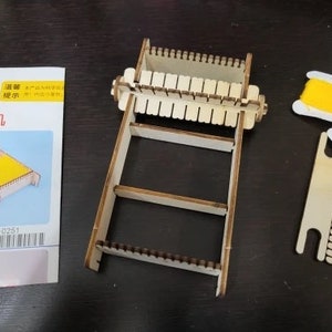 May include: A wooden loom with a yellow and red spool of thread. The loom has a wooden frame with a series of slots for weaving. The spool of thread is attached to a small wooden stand. The text on the packaging reads "织布机" which translates to "loom" in English.