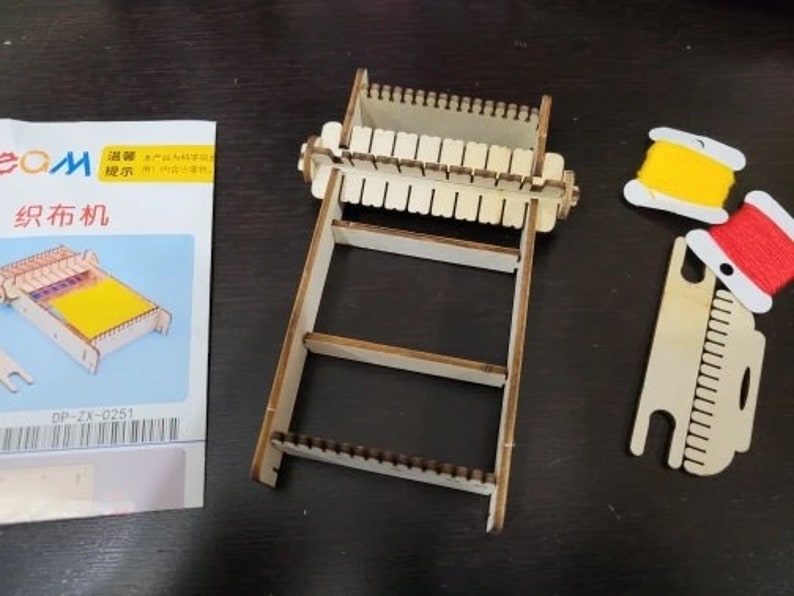 May include: A wooden loom with a yellow and red spool of thread. The loom has a wooden frame with a series of slots for weaving. The spool of thread is attached to a small wooden stand. The text on the packaging reads "织布机" which translates to "loom" in English.