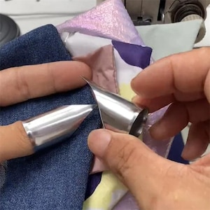 May include: Close-up of silver metal thimbles with pointed tips. The thimbles are being used on denim fabric and other colorful fabrics. The thimbles are designed to protect fingers while sewing or crafting.