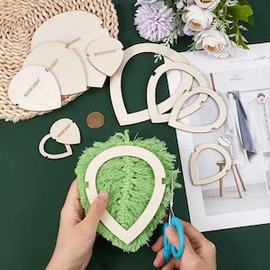 May include: A collection of unfinished wooden craft supplies, including heart and teardrop shapes, and a circular frame. A pair of hands holds a green macrame piece with a wooden frame, and blue scissors are used to trim it.