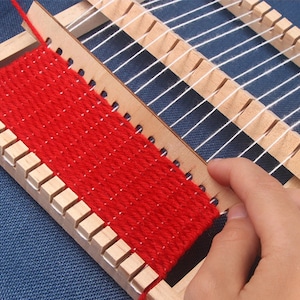May include: A wooden loom with red yarn woven into a small tapestry. The loom has a white warp thread and a wooden shuttle.