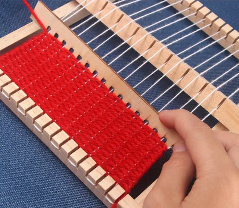 May include: A wooden loom with red yarn woven into a small tapestry. The loom has a white warp thread and a wooden shuttle.