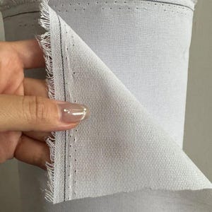 Puede incluir: Un rollo de tela gris claro con una textura tejida visible. El borde del material está deshilachado y presenta una fina línea vertical oscura paralela al orillo, marcado por una serie de pequeños orificios espaciados uniformemente.