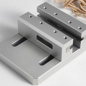 May include: A silver metal vise with a rectangular base and two parallel clamping jaws. The jaws have multiple screw holes. The vise is designed for holding objects securely. The background is a light colour.