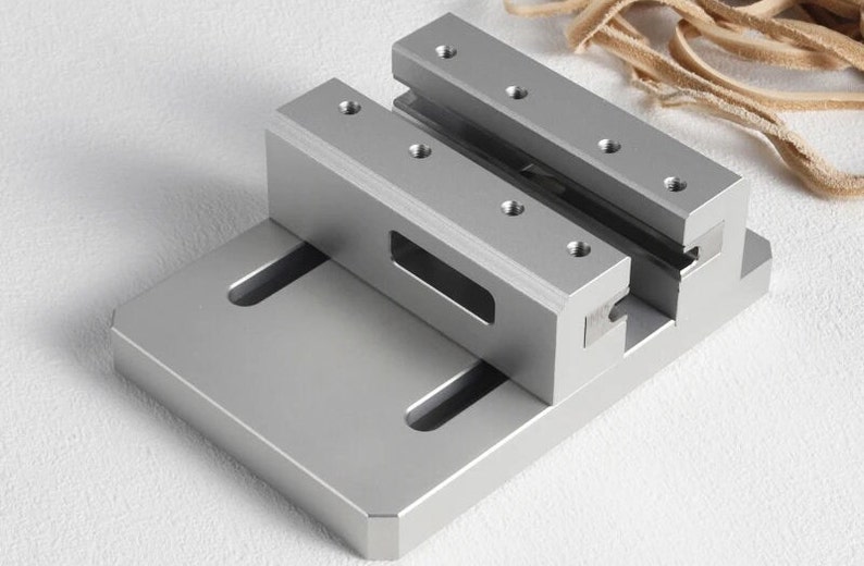 May include: A silver metal vise with a rectangular base and two parallel clamping jaws. The jaws have multiple screw holes. The vise is designed for holding objects securely. The background is a light colour.
