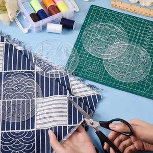 Op de afbeelding: Een knutselscène met stof, schaar en naaimaterialen. Een blauw-wit patroonstof wordt met een schaar geknipt. Transparante sjablonen, klosjes garen en een snijmat zijn ook zichtbaar, wat wijst op een naai- of knutselproject.