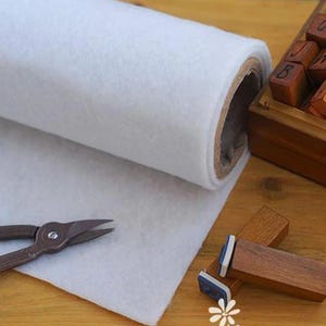 Può includere: Un grande rotolo di feltro bianco posizionato accanto a un paio di forbici in metallo marrone scuro e diversi blocchi di legno, inclusa una piccola scatola di legno contenente diversi timbri quadrati in legno con lettere incise sulle superfici.