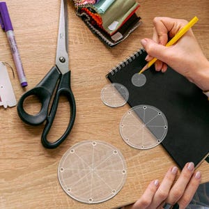 Peut inclure: Un espace de travail artisanal avec des gabarits circulaires en acrylique transparent, des ciseaux, un marqueur violet et des échantillons de tissu. Une personne utilise un crayon jaune pour dessiner sur un carnet noir. Les gabarits ont des sections et des trous marqués.