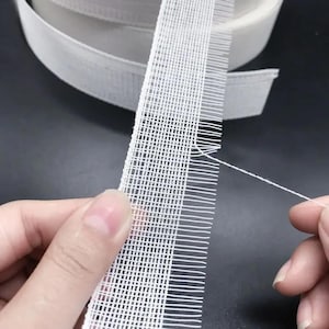 May include: A close-up of white woven fabric tape with a grid-like pattern. The tape is held by two hands, with a loose thread extending from the edge. A roll of the same tape is visible in the background, suggesting a crafting or sewing supply.