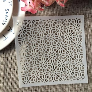 May include: A square, semi-transparent plastic stencil featuring a repeating geometric pattern of interconnected circles and lines. The design consists of a grid of floral-like shapes with thin, cut-out lines that form a symmetrical, lattice-like structure.